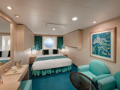 Eine gemütliche Aussenkabine an Bord der MSC Musica mit einem großen Doppelbett, zwei Nachttischen und einem runden Porthole, das einen weiten Blick auf das blaue Meer und den Himmel bietet. Ein türkisfarbener Teppich und passende Möbel setzen Farbakzente.