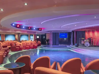 Eine geräumige Lounge an Bord der MSC Musica mit orangefarbenen Sesseln, einem blauen Tanzbereich und einer beleuchteten Bühne mit Schlagzeug.