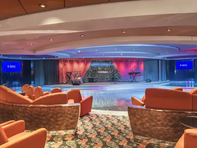 Die Crystal Lounge an Bord der MSC Musica mit einer Bühne, beleuchteter Kulisse und Musikinstrumenten. Eine Tanzfläche wird von geschwungenen, orangen Sofas umrahmt. Zwei Bildschirme zeigen das MSC-Logo.