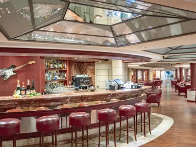 L'Enoteca Wine Bar an Bord der MSC Musica: Geschwungene Theke mit roten Barhockern, Flaschenregale, Kaffeemaschine, Deko-Gitarre. Im Hintergrund gemütliche rote Ledersessel.