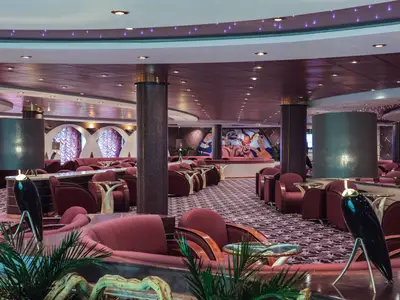 Die weitläufige Il Tucano Lounge an Bord der MSC Musica, ausgestattet mit opulenten roten Samtsesseln, runden Tischen und einem gemusterten Teppich. Zwei grosse Lampen und eine kunstvolle Wandmalerei prägen den Raum.