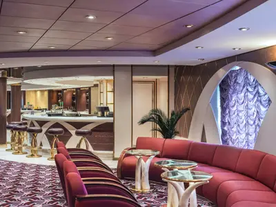 Ansicht der Il Tucano Lounge an Bord der MSC Musica. Eine geschwungene Bar mit Hochhockern und roten Samtsofas sind zu sehen. Im Hintergrund eine lila-beleuchtete Wanddekoration.