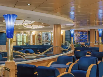 Blick in die elegante Blu Velvet Bar an Bord der MSC Musica. Blaue Sessel, goldene Akzente und besondere blaue Tulpen-Lampen schaffen eine gemütliche Atmosphäre. Boutiquen im Hintergrund.