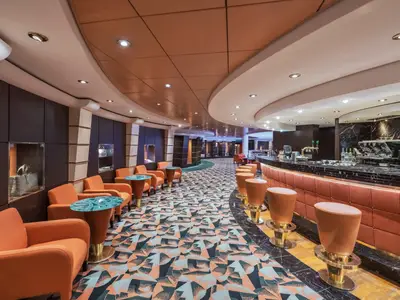 Die stilvolle The Golden Bar an Bord der MSC Musica. Der langgezogene Barbereich mit Hockern und einladenden Sesseln im warmen Orangeton sind zu sehen, dazu ein geometrischer Teppich.