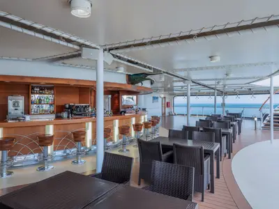 Weitwinkelaufnahme der überdachten Blu Marlin Bar an Bord der MSC Musica mit Holztheke, Barhockern, Tischen und Stühlen. Im Hintergrund ist das Meer sichtbar. Über der Bar hängt eine große Fischskulptur.