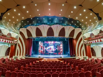 Weitwinkelaufnahme des Teatro La Scala an Bord der MSC Musica mit Reihen von roten Samtsitzen, die auf eine beleuchtete Bühne mit grosser LED-Leinwand blicken. Die Innenarchitektur ist reich verziert mit muschelförmigen Lichtelementen und geschwungenen Mustern.