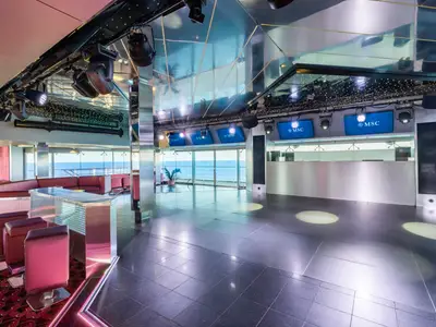 Blick in die moderne Q32 Disco an Bord der MSC Musica. Der große Raum umfasst eine leere Tanzfläche, eine Bar mit roten Hockern und rote Lounge-Sitzbereiche, alles mit großen Fenstern zum Meer. Die Decke ist verspiegelt, und über der Bar sind Bildschirme mit dem MSC-Logo.