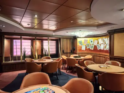 Elegantes Kartenzimmer an Bord der MSC Musica mit mehreren runden Tischen und bequemen Ledersesseln. Eine Wand ist mit großen Spielkarten dekoriert. Durch Fenster scheint pinkfarbenes Licht herein.