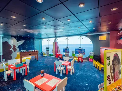 Ein lebhafter Kinderbereich an Bord der MSC Musica mit einem Weltraum-Wandgemälde, einem Kicker, kleinen Tischen mit Stühlen, interaktiven Bildschirmen und einem runden Fenster mit Meerblick. Der Boden ist mit einem blauen Teppich bedeckt, der an Sterne erinnert.