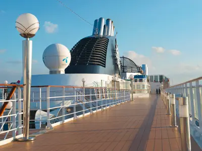 Blick auf das Sonnendeck der MSC Musica mit charakteristischem Schornstein, Telenor-Satellitenkuppel, Holzdeckweg und weißen Geländern unter blauem Himmel.