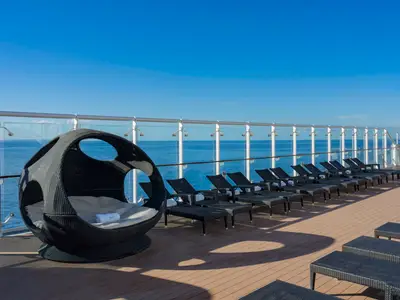 Ein weitläufiges Sonnendeck der MSC Musica an einem sonnigen Tag. Im Vordergrund steht eine schwarze, ovale Rattan-Tagesliege mit Kissen. Dahinter reihen sich mehrere schwarze Sonnenliegen entlang einer Glasbrüstung, die den Blick auf das strahlend blaue Meer freigibt. Der Himmel ist wolkenlos.