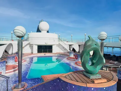 Ein sonniger Blick auf den La Spiaggia Poolbereich an Bord der MSC Musica. Ein türkisfarbenes Becken und ein blau-weiß gefliester Whirlpool sind von Holzdecks und Liegestühlen umgeben. Eine große bronzene Skulptur steht prominent im Vordergrund, mit dem blauen Meer im Hintergrund unter einem klaren Himmel.