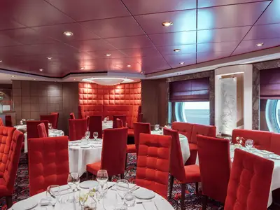 Innenansicht des eleganten Restaurants Belle Époque an Bord der MSC Musica. Rote, gepolsterte Stühle und Sofas um weiss gedeckte Tische. Runde Bullaugen mit Blick auf das blaue Meer.