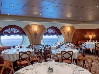 Elegantes Restaurant L'Oleandro an Bord der MSC Musica. Runde Tische mit weißen Decken, Holzstühle und rote Sitzbänke bieten Meerblick durch große Bullaugenfenster. Dezente Beleuchtung schafft eine ruhige Atmosphäre.