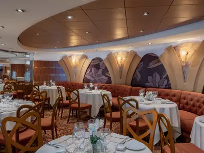 Ein elegantes Restaurant L'Oleandro an Bord der MSC Musica. Gedeckte Tische, Holzstühle und rote Sitzbänke sind sichtbar. Florale Wanddekorationen und warme Beleuchtung schaffen ein einladendes Ambiente. Der Raum ist unbesetzt.