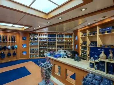 Der helle Shop der MSC Musica zeigt Regale voller Souvenirs, darunter Plüschtiere, Mützen und Parfums, sowie eine Kassentheke. Der Boden ist mit blau-orangem Teppich ausgelegt.