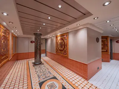Heller Flur im MSC Aurea Spa der MSC Musica, verziert mit kunstvollen Mosaikwänden, einer Mosaiksäule und gemusterten Fliesenböden. Schilder leiten zu Sauna und Dampfbad.