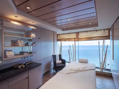 Ein moderner Spa-Behandlungsraum an Bord der MSC Musica mit einer weissen Massageliege, Handtüchern, Wellnessprodukten und grossen Fenstern mit Meerblick. Bambusrohre stehen dekorativ vor den Fenstern, die den weiten Ozean zeigen.