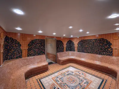 Ein breiter Blick in den Thermalbereich des MSC Aurea Spa an Bord der MSC Musica. Der Raum ist mit orangefarbenen und schwarzen Mosaikfliesen dekoriert. Grosse, geschwungene, geflieste Baenke laden zum Entspannen ein. In der Mitte des Raumes befindet sich ein kunstvolles Bodenmosaik. Warme Deckenlichter erhellen den Raum.