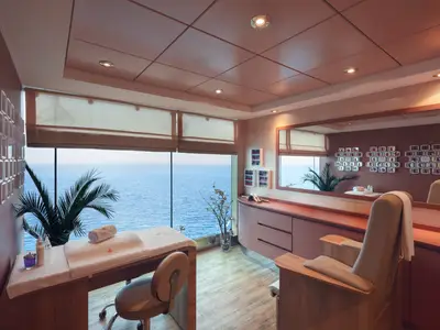 Ein helles Nagelstudio im MSC Aurea Spa der MSC Musica mit großen Fenstern und weitem Blick auf das blaue Meer. Ein Behandlungsbett und ein Stuhl sind zu sehen, sowie Regale mit Nagellacken an der Wand.