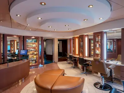 Ein heller und moderner Haarsalon an Bord der MSC Musica mit mehreren Styling-Stühlen, großen Spiegeln, beleuchteten Produktregalen und einer geschwungenen Rezeptionstheke. Der Boden ist gefliest, die Wände sind in warmen Brauntönen gehalten.