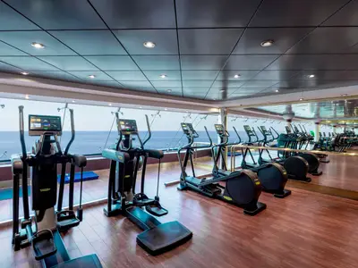 Ein modernes Fitnessstudio an Bord der MSC Musica mit Reihen von Technogym-Crosstrainern, die auf große Fenster mit Blick auf das Meer ausgerichtet sind. Der Raum hat dunklen Holzboden, helle Deckenpaneele und Spiegel an einer Wand. Keine Personen sichtbar.