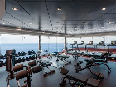 Ein geräumiges Fitnessstudio an Bord der MSC Musica mit Meerblick. Zahlreiche Technogym-Geräte wie Laufbänder und Hantelbänke sind zu sehen, mit dem weiten blauen Meer im Hintergrund.