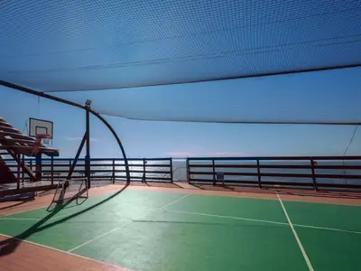 Sportplatz an Bord der MSC Musica. Man sieht einen Basketballkorb, ein grünes Spielfeld, umgeben von einem Netz und Holzreling, mit Blick auf das blaue Meer.