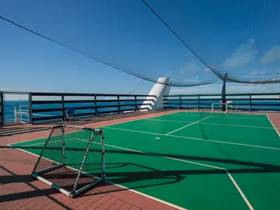 Sportfeld auf der MSC Musica: Grüne Fläche mit weißen Linien und zwei Toren. Schutznetz oben, dahinter das blaue Meer und der Himmel.