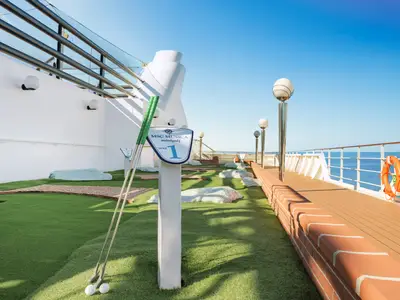 Minigolfplatz an Deck der MSC Musica. Grüne Bahnen, Hindernisse, Loch 1 und Minigolfschläger. Rechts ein Holzdeck mit Rettungsring und weitem Meer unter blauem Himmel.