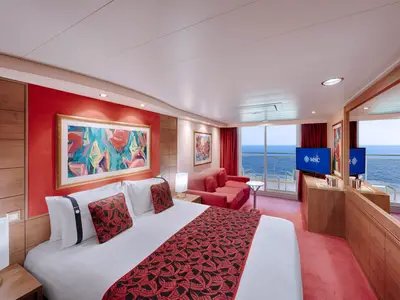 Geräumige Premium Suite Aurea an Bord der MSC Musica. Sie zeigt ein großes Doppelbett, eine rote Couch, einen Fernseher und einen Balkon mit Blick auf das blaue Meer.