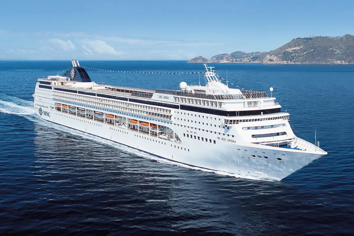 Die MSC Opera, ein großes Kreuzfahrtschiff, fährt auf dem tiefblauen Meer. Im Hintergrund sind grüne Inseln und eine Küstenlinie unter einem hellblauen Himmel zu sehen. Am Heck zieht das Schiff eine deutliche Schaumspur.