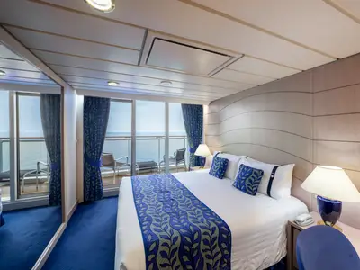 Moderne Balkonkabine an Bord der MSC Opera mit Doppelbett, blauer Bettwaesche und Meerblick. Glastuer zum Balkon mit Stuehlen.