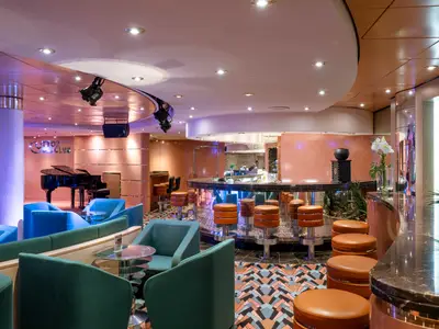 Der elegante Cotton Club an Bord der MSC Opera zeigt eine moderne Bar mit Hockern und Flügel. Komfortable Sitzgruppen in Blau und Grün laden ein. Warme Beleuchtung prägt das gemütliche Abendambiente.