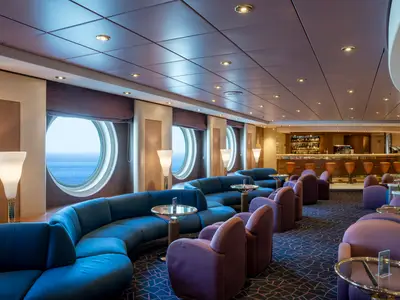 Geräumige Lounge an Bord der MSC Opera mit blauen und lila Samtsofas, runden Bullaugen mit Meerblick und einer Bar im Hintergrund. Warme Beleuchtung durch Deckenstrahler und Stehlampen.