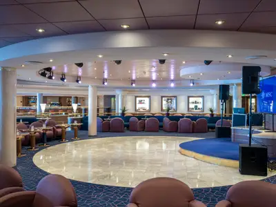 Die Piazza di Spagna Bar an Bord der MSC Opera zeigt eine helle Lounge mit Bar, violetter Bestuhlung und einem zentralen Tanzbereich mit Bühne. Ein Bildschirm zeigt das MSC Logo.