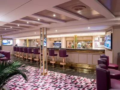 Bar an Bord der MSC Opera: lange Theke, lila Barhocker, viele Getränke. Zwei Bildschirme. Gemusterter Teppich und Palme. Elegantes Interieur.