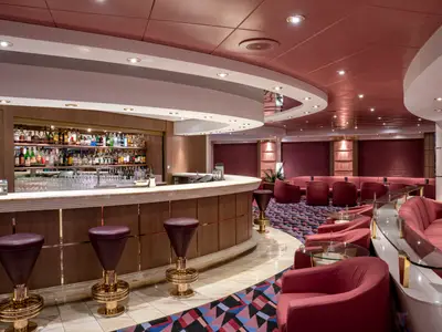 Blick in die schicke Bar 'La Cabala' an Bord der MSC Opera, mit geschwungenem Tresen, hohen Hockern und zahlreichen roten Sitzmöbeln. Rote Decke, Boden mit geometrischem Teppich.