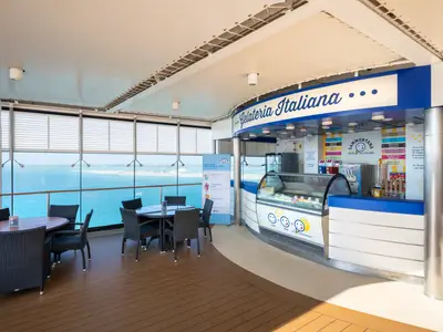 Eine helle Gelateria Italiana an Bord der MSC Opera mit einer Theke voller Eissorten und Tischen. Grosse Fenster bieten einen weiten Blick auf das blaue Meer und die Kueste.