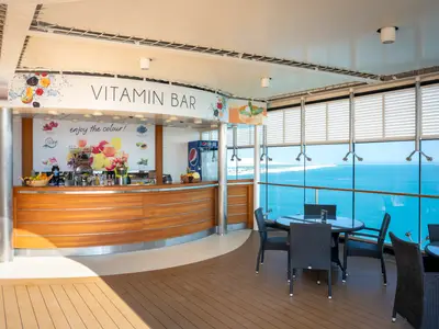 Die Vitamin Bar an Bord der MSC Opera. Halbrunde Holztheke mit frischen Früchten. Sitzbereich mit Tischen und Stühlen bietet weiten Meerblick auf eine Küste.