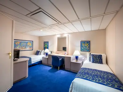 Die helle Innenkabine an Bord der MSC Opera zeigt zwei Einzelbetten mit blauen Mustern, einen blauen Teppich und einen Schreibtisch mit Stuhl in der Mitte. Farbefrohe Kunstwerke schmücken die Wände.