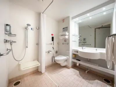 Ein helles, barrierefreies Badezimmer an Bord der MSC Opera, ausgestattet mit Duschsitz, Haltegriffen, Duschbrause, WC, Waschbecken und Spiegel.