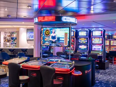 Casino-Bereich "Med Pearl" auf der MSC Opera mit Spielautomaten und digitalen Roulettetischen. Ein Bildschirm zeigt eine Live-Dealerin; im Hintergrund eine Bar.