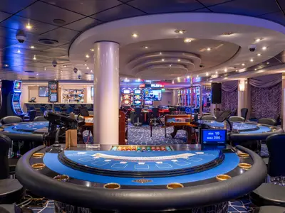Das Med Pearl Casino an Bord der MSC Opera mit blauen Spieltischen im Vordergrund. Leuchtende Slot Machines und eine Bar im Hintergrund. Moderne Beleuchtung.