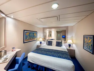 Innenkabine der MSC Opera mit Doppelbett, blauen Textilien und farbenfrohen Kunstwerken an den Waenden. Ein Schreibtischbereich und Spiegel erweitern den Raum optisch.