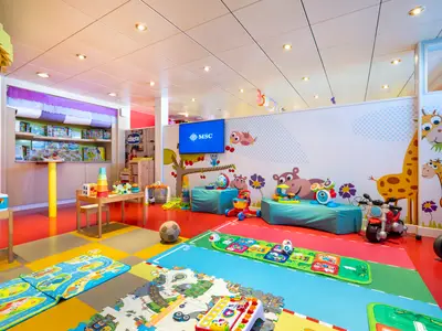 Im Baby Club der MSC Opera: bunte Spielzeuge, Kindermöbel und weiche Spielmatten. Wände mit lustigen Tiermotiven und MSC-Logo auf dem Bildschirm.