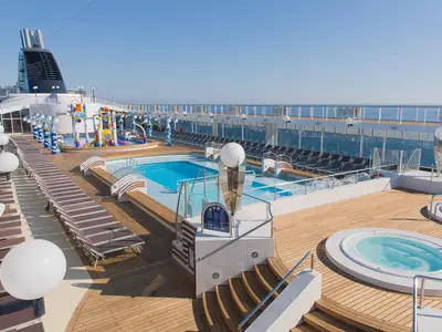 Das sonnige Pooldeck der MSC Opera mit dem Hauptpool, dem bunten Doremi Spray Park und zwei Whirlpools. Liegestühle und das offene Meer unter blauem Himmel sind zu sehen.