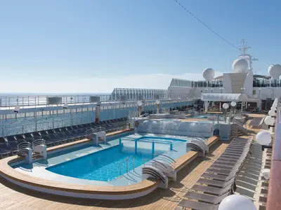 Ein sonniges Deck mit großem Pool und Whirlpools an Bord der MSC Opera. Reihen von Liegestühlen säumen die Holzterrassen, im Hintergrund ist das Meer zu sehen.