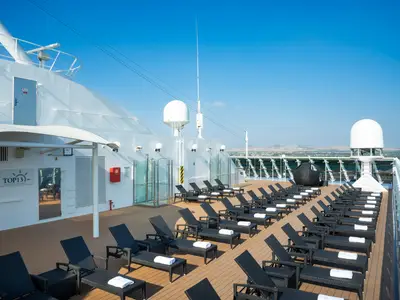 Das Top 13 Exclusive Solarium an Bord der MSC Opera zeigt eine Reihe von schwarzen Liegestühlen mit weißen Handtüchern auf einem Holzdeck unter strahlend blauem Himmel. Im Hintergrund sind Berge und das Meer zu sehen.