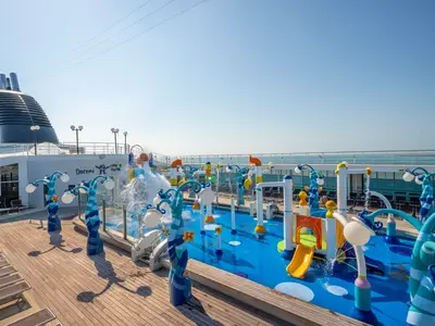 Der farbenfrohe Doremi Spray Park an Bord der MSC Opera bei Tag. Der Wasserpark umfasst Rutschen und spritzende Fontänen. Links der Schornstein des Schiffes, rechts das weite Meer.